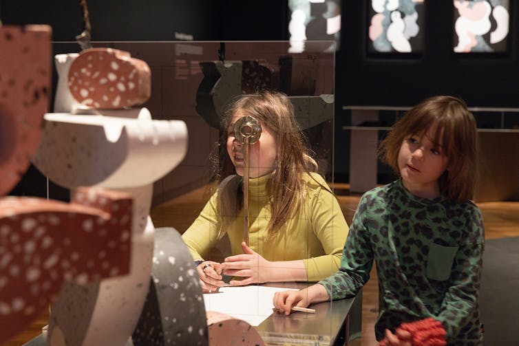 Two children look at a sculpture; one is drawing it.