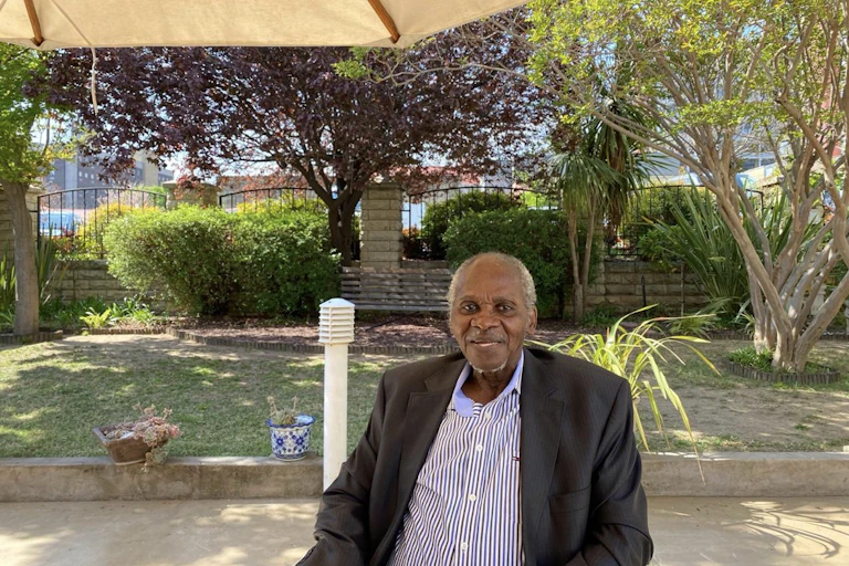 An elderly African man with grey hair smiles for a photo in a garden.