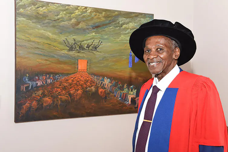 An elderly African man in academic robes smiles as he poses next to a dramatic painting featuring cattle, humans and god-like figures.