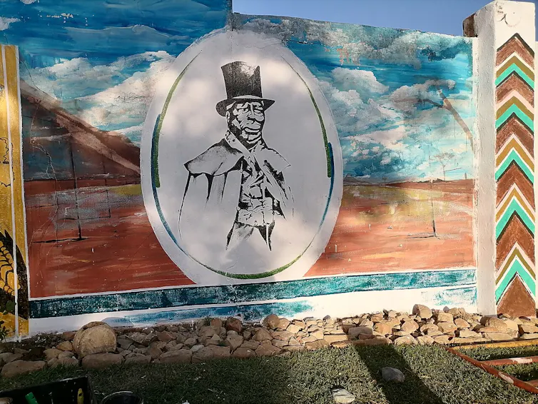 A wall painting of land and sky featuring an oval portrait of a smartly dressed older African man.