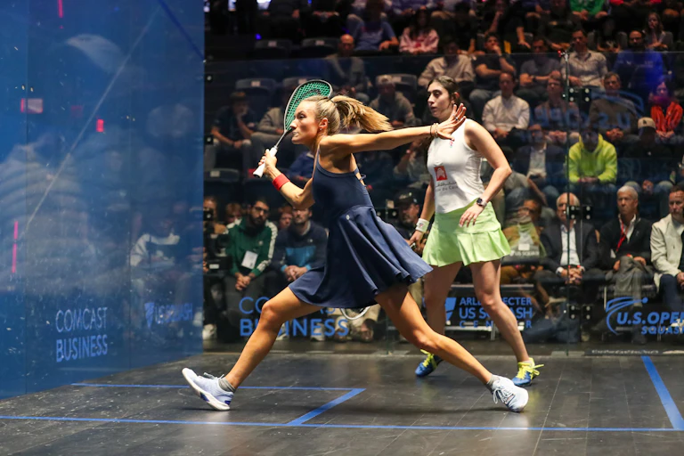 Two women compete on squash court with crowd watching