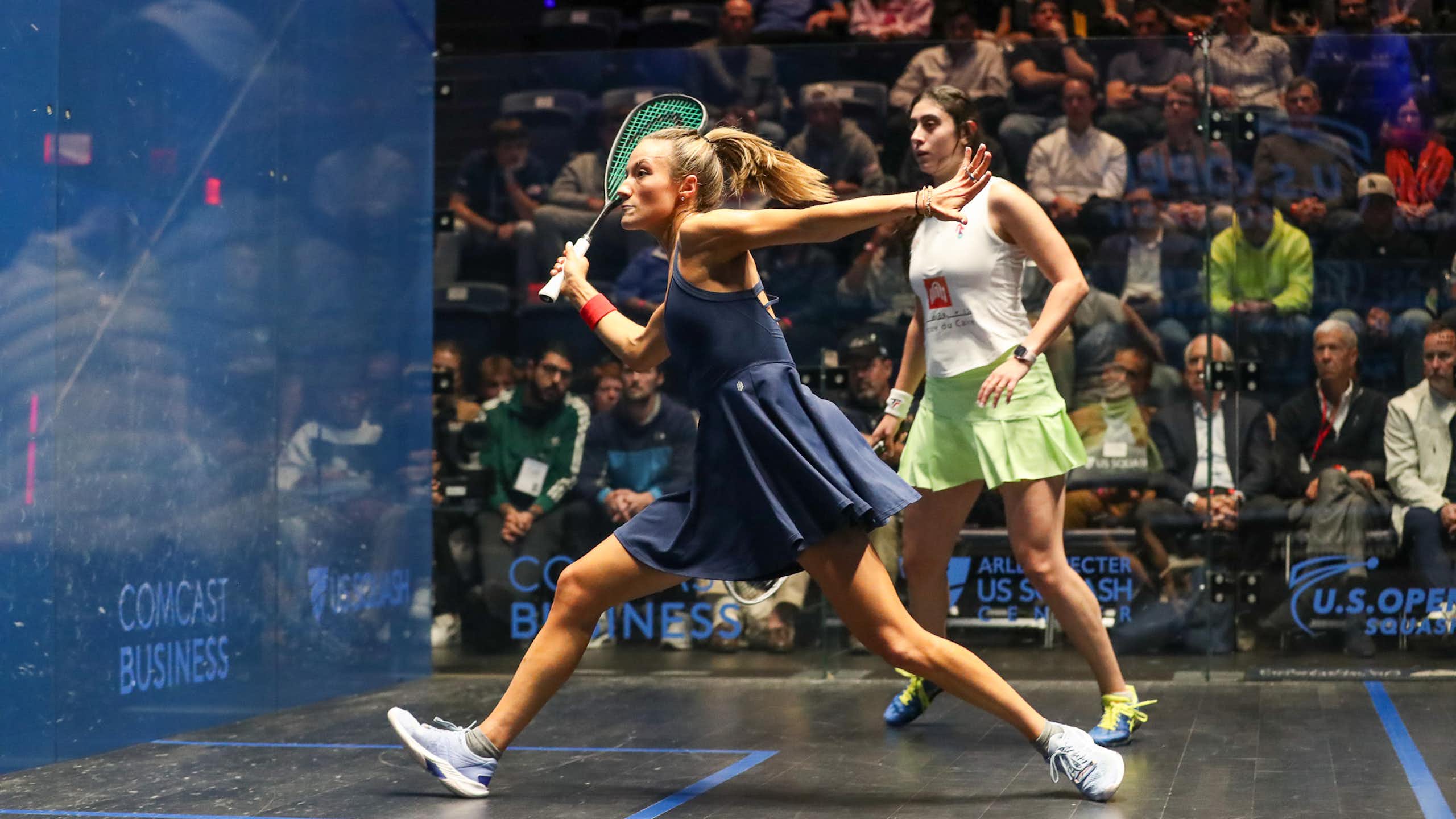 Two women compete on squash court with crowd watching