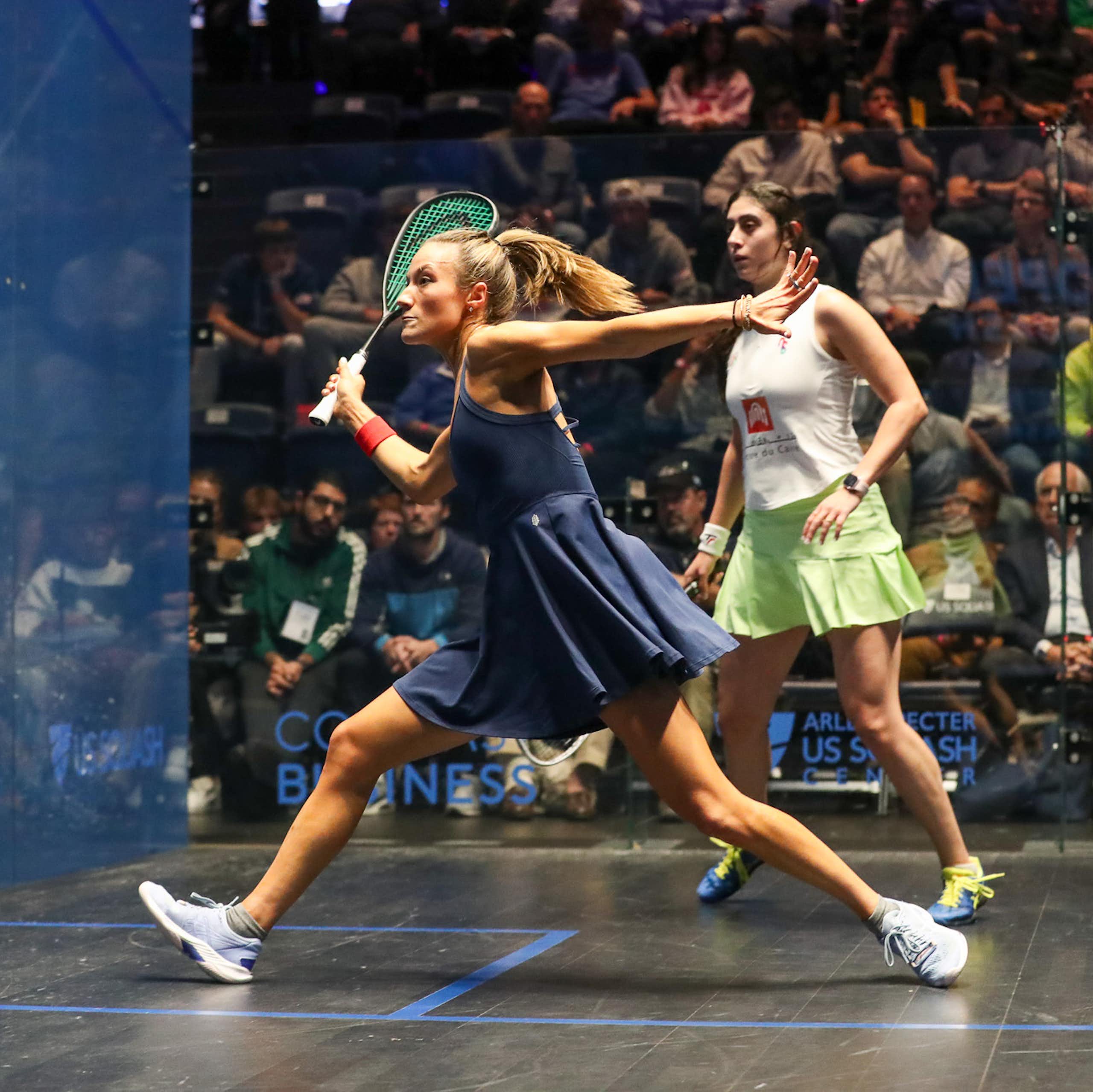 Two women compete on squash court with crowd watching