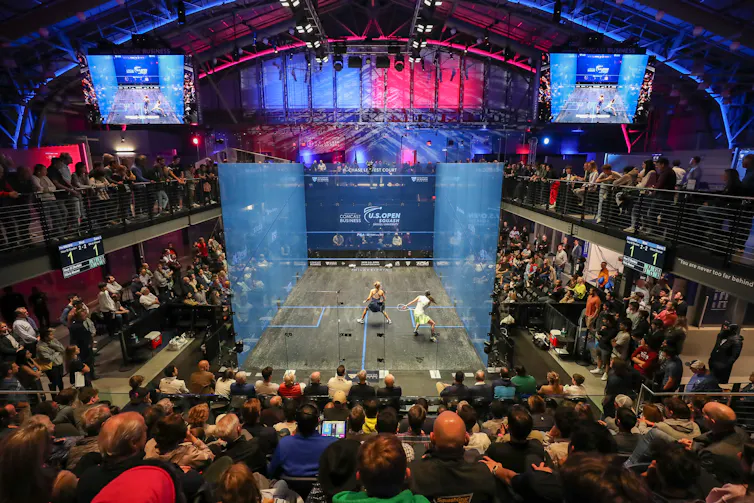 Two women athletes compete on squash court with four transparent walls surrounded by onlookers