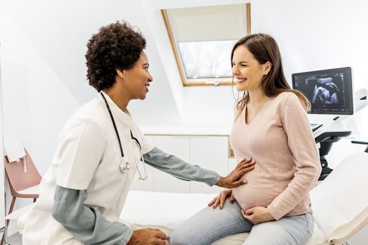Smiling pregnant woman talking with a female doctor, with an ultrasound screen behind them.