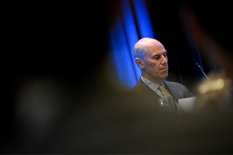 A man dressed in suit and tie sits at a table on a stage.