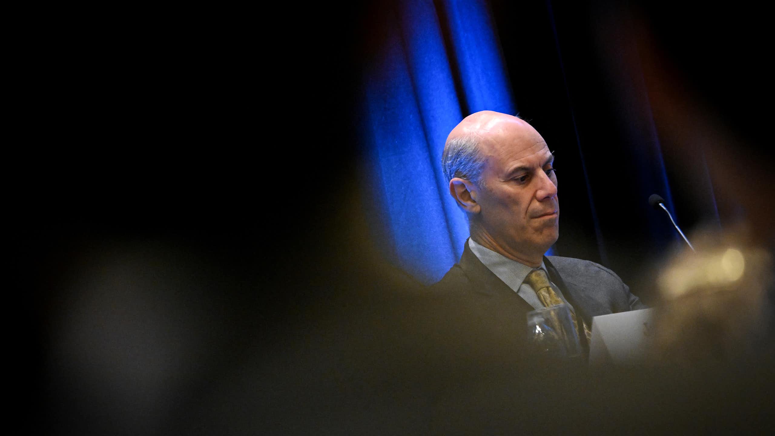 A man dressed in suit and tie sits at a table on a stage.