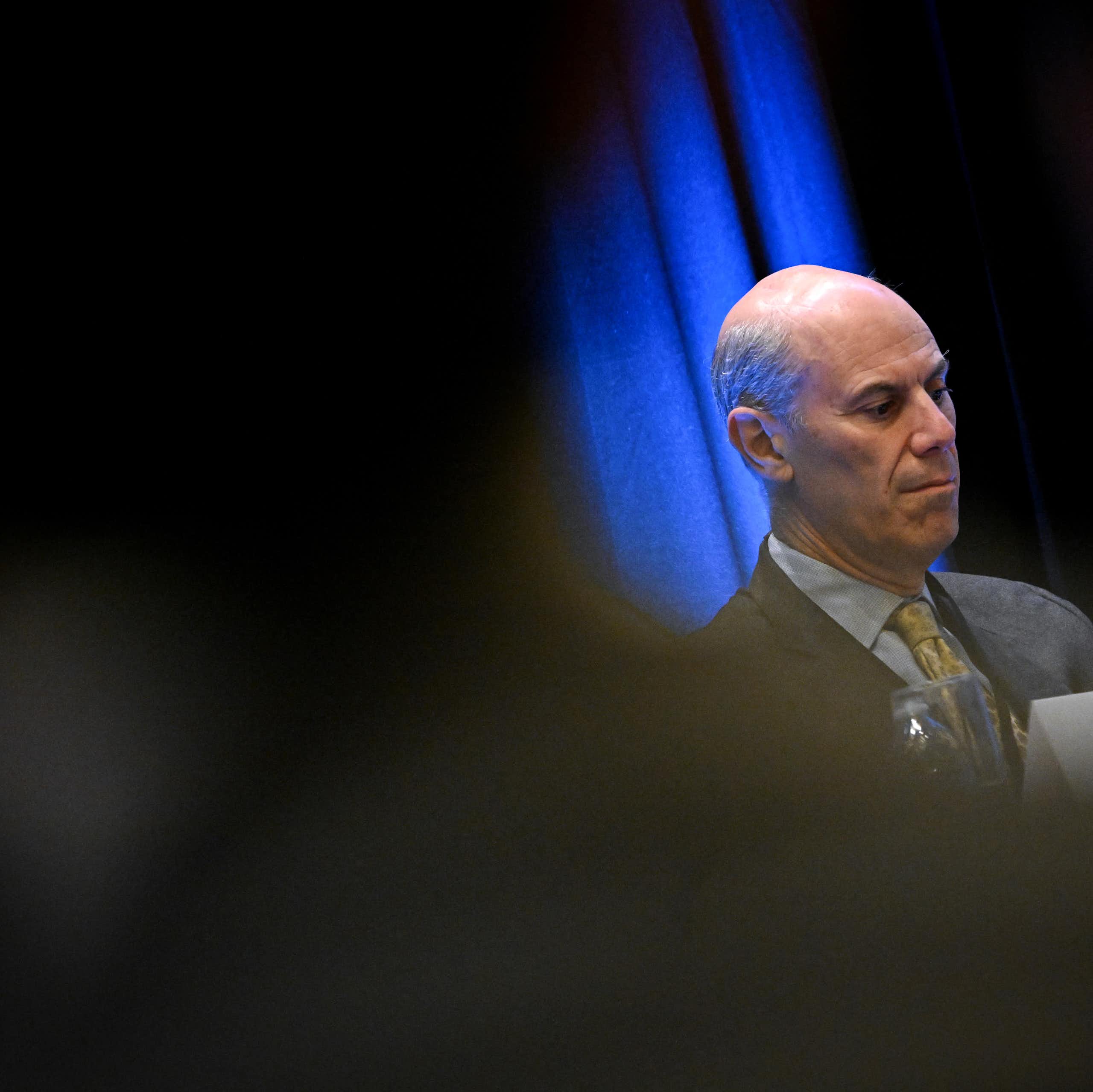 A man dressed in suit and tie sits at a table on a stage.