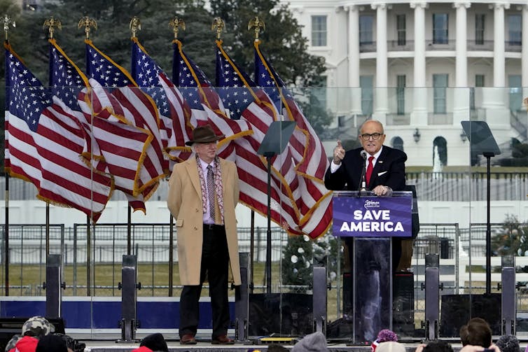 A man in a suit and coat stands next to another man in front of a lectern, with American flags and the White House in the background.