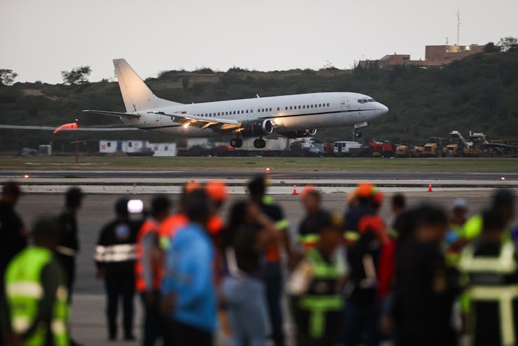 An airplane lands on a runway as people watch in the foreground.