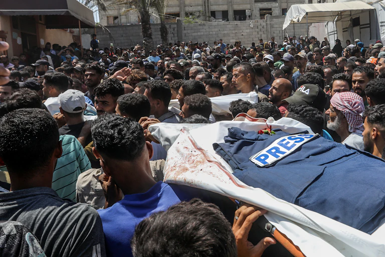 Th body of a person with a 'press' jacket on top is carried by mourners through the streets.