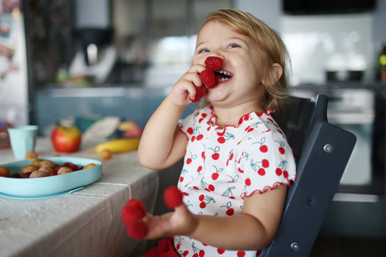 Smiling toddler eats raspberries off her fingers.