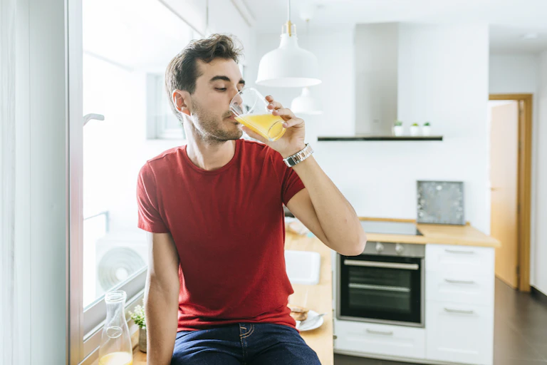 A man sitting in his kitchen drinking an orange drink.