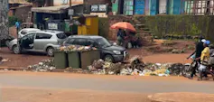 Trois grandes poubelles en plastique sur le bord d'une rue débordant de déchets éparpillés le long de la route.