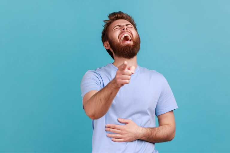 A young man against a blue wall, pointing and laughing mockingly at the camera