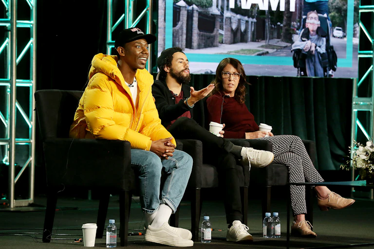 Two young men, one wearing a yellow jacket, are seated with a young woman and talking.