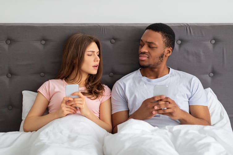 You couple with smartphones sitting in bed with phones and looking at each other with suspicion.