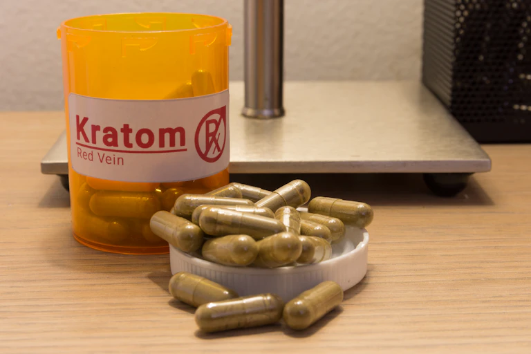 A yellow pill bottle labeled 'kratom' sits next to its cap full of green capsules.