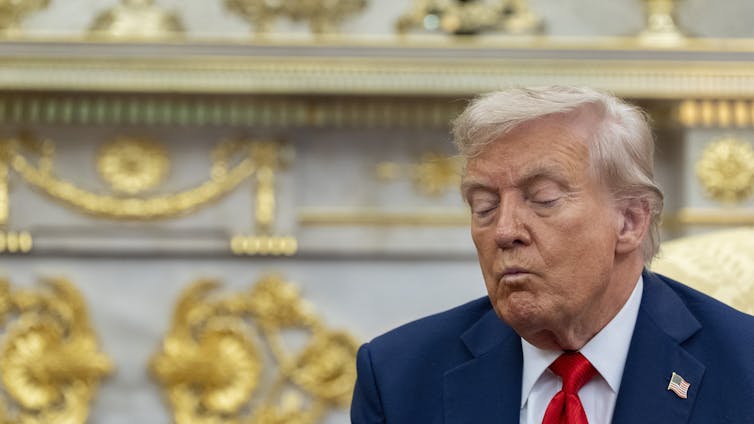 A man with orange-ish white hair in a blue suit and red tie sits with his eyes closed in an ornate office with gilded ornaments behind him.