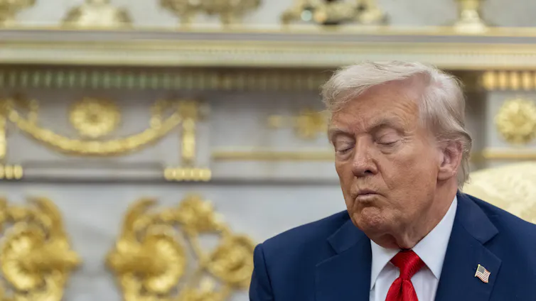 A man with orange-ish white hair in a blue suit and red tie sits with his eyes closed in an ornate office with gilded ornaments behind him.