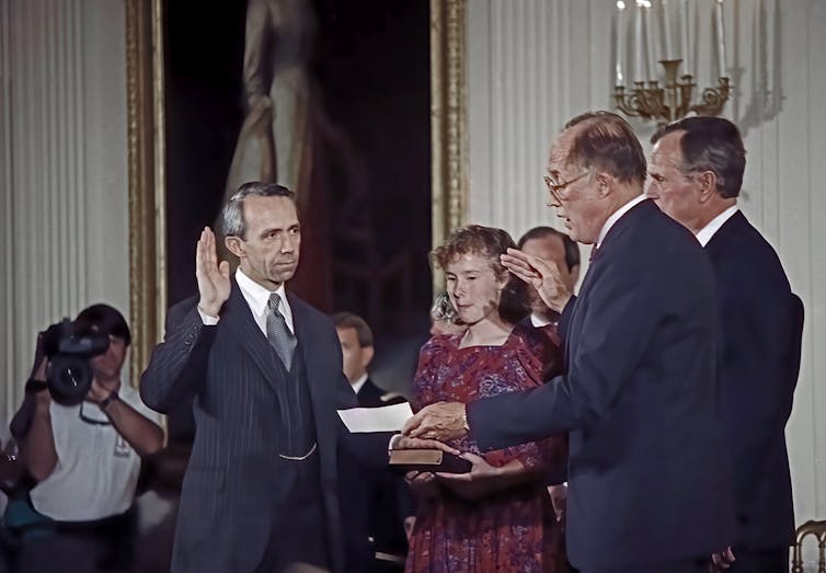 A man in a suit and tie places his left hand on a bible and raises his right hand as he receives an oath.
