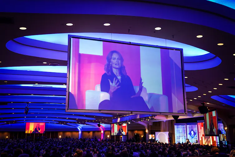 A woman, her image projected on a big screen, speaks to an audience.