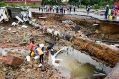Dozens of people stand next to a long road that has collapsed while others try to collect water that is leaking out of a pipe