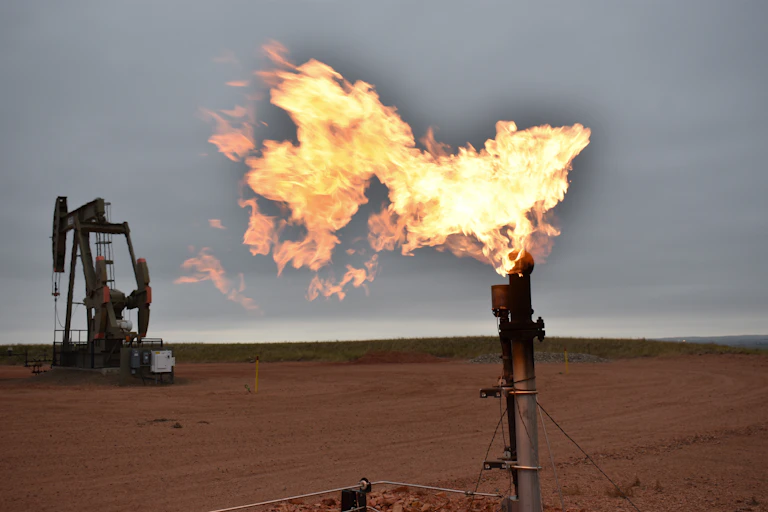 A pump jack above an oil well sits in the background of a large flame coming out of a pipe.
