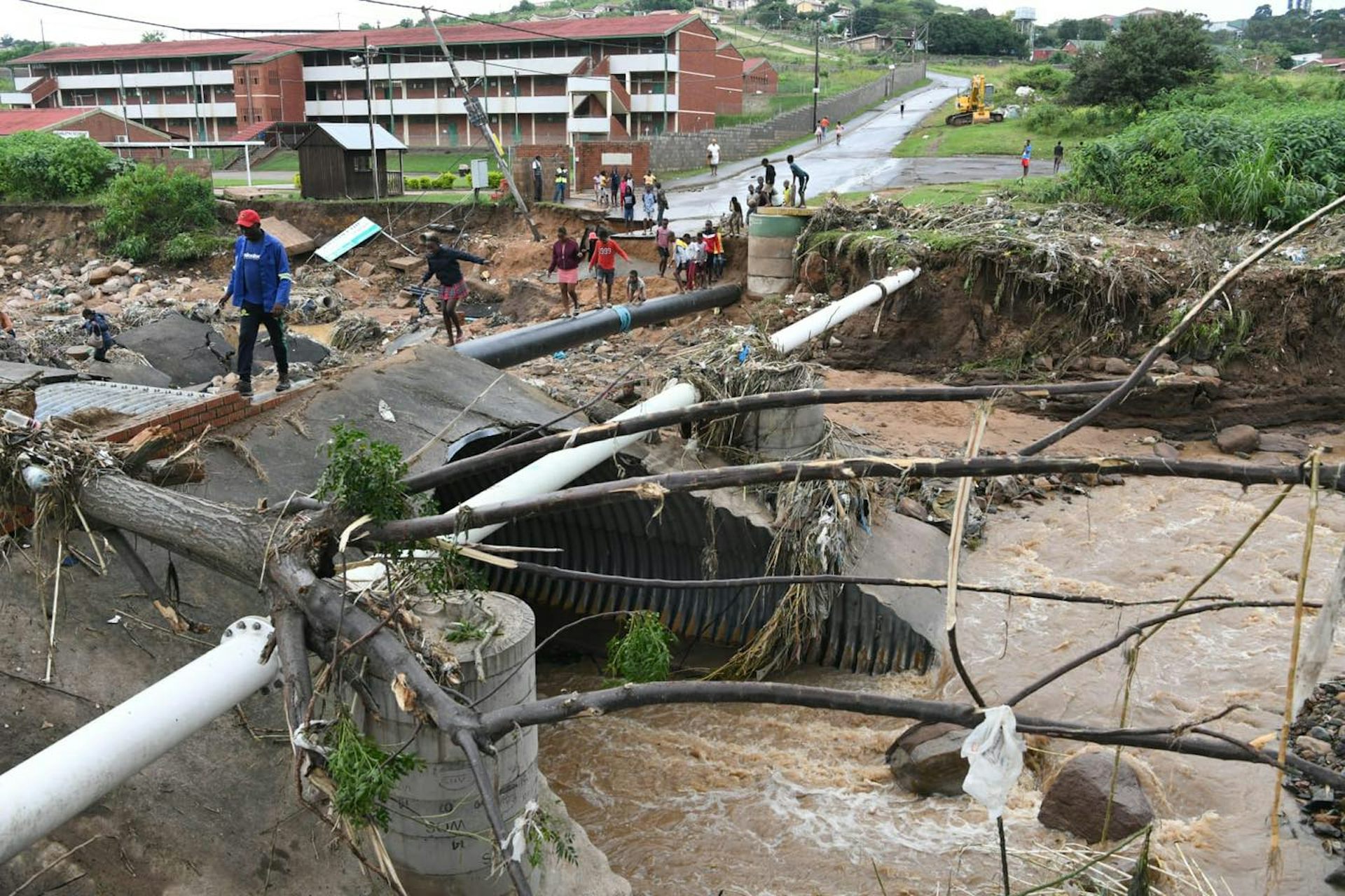 A road that has been partly washed away by a river, exposing pipes which people are walking on