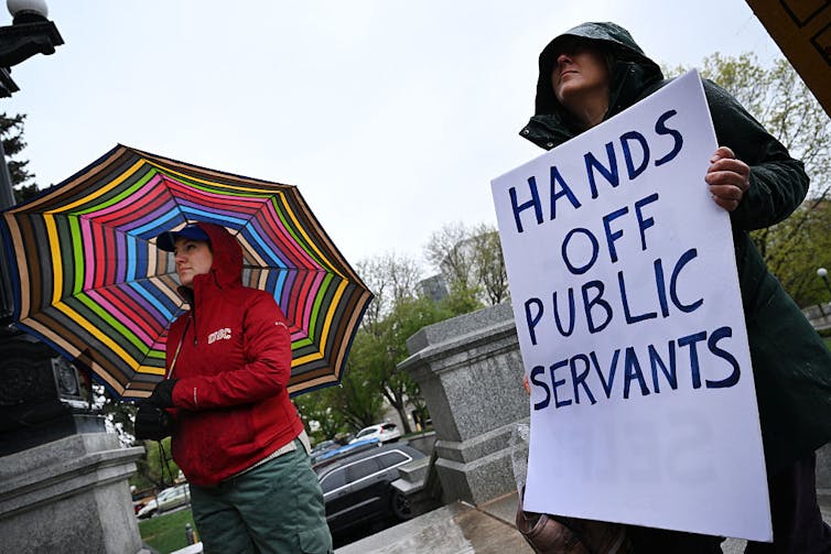 Two people in raincoats stand outside, with one holding a sign that says 'Hands off public servants.'