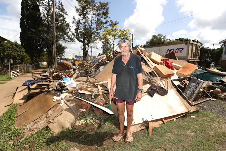 A man stands in front of flood-soaked debris from his home.