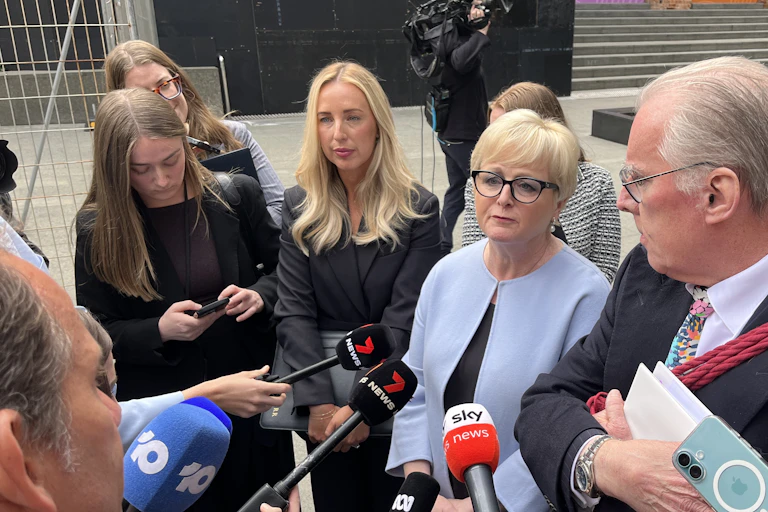 A woman stands surrounded by reporters pointing microphones towards her