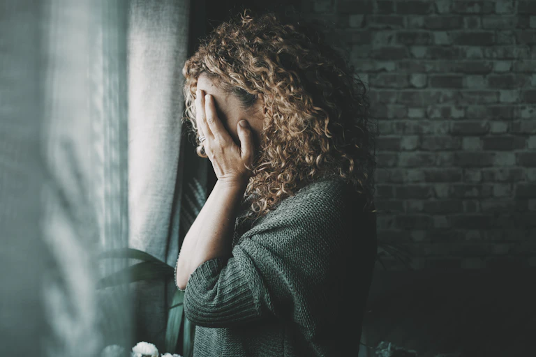 A woman with her face in her hand standing by a window at home.