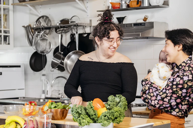 A smiling trans person in a kitchen smiles at another holding a baby.