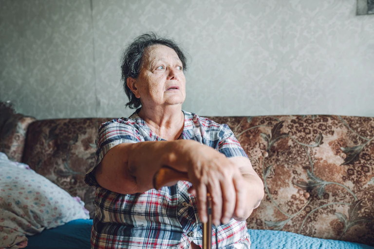 A woman sitting in a somewhat dilapidated room, with her hand over a walking stick.