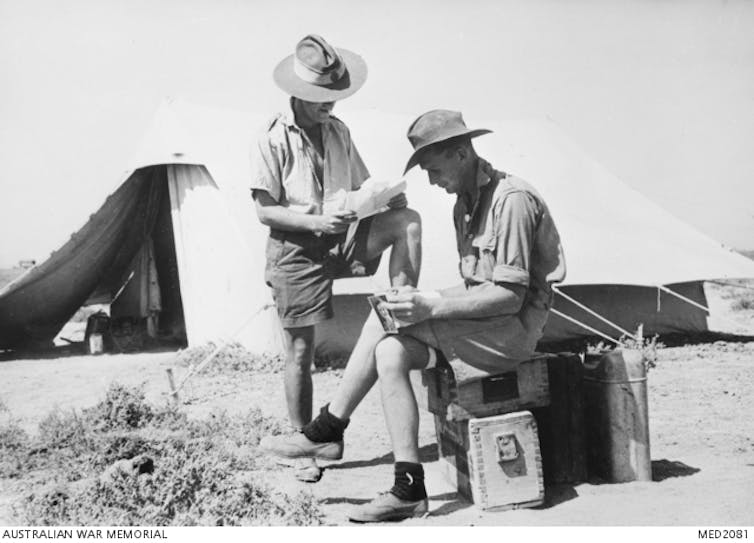 Two men read letters outside tents.