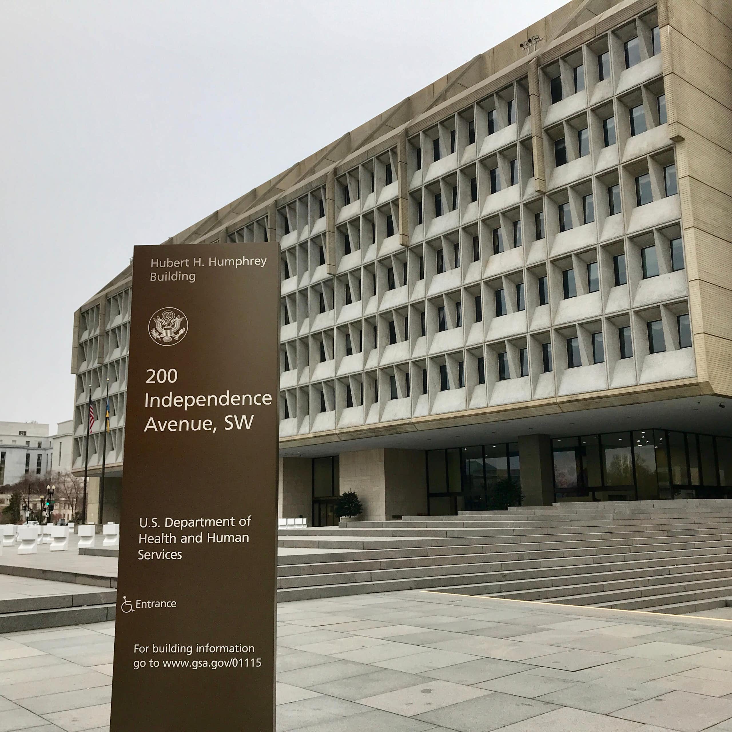 A vertical sign with the words "Hubert H. Humphrey Building" and "U.S. Department of Health and Human Services" stands on a gray stone plaza, with a large, low grayish building behind it, beneath an overcast sky.