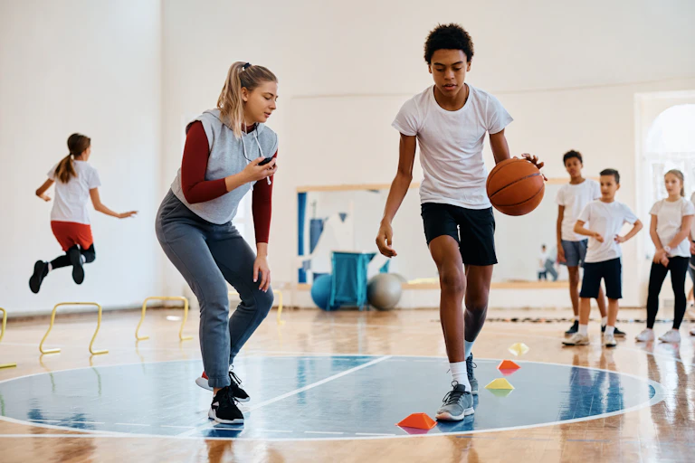 A boy dribbles a basketball around a cone on the floor while their female teacher supervises.