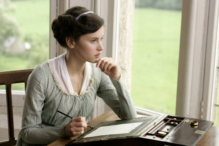 A young Regency period woman sitting at a writing desk looking out of the window.