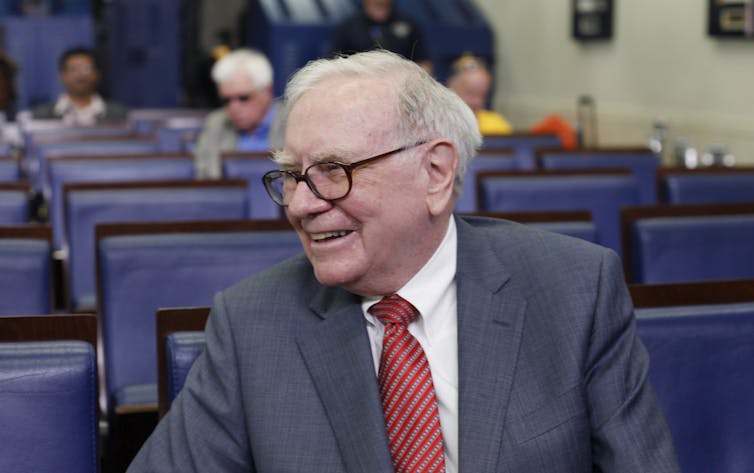 A man with white hair and brown glasses, dressed in a suit and tie, smiles as he sits on a blue chair.