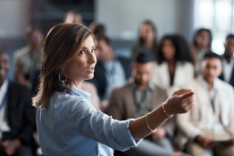 A brunette woman in a blue button-up shirt gestures as she speaks to a room of people.