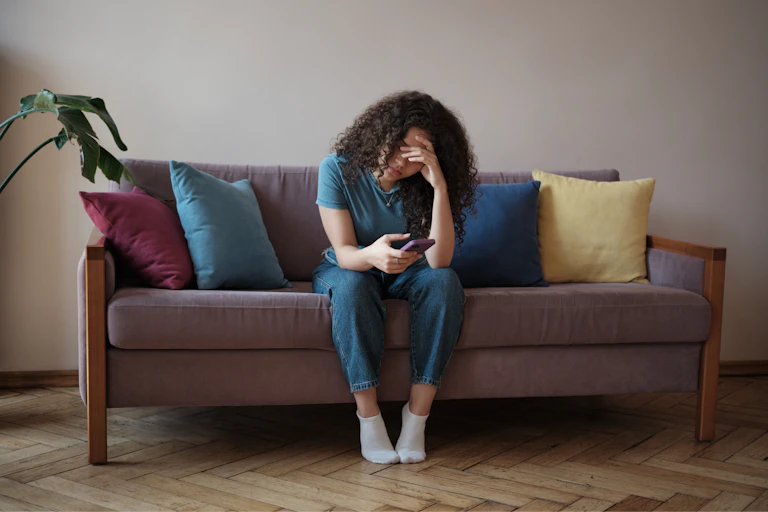 A teen girl sits on a couch, bracing her forehead with one hand and looking at her cell phone.
