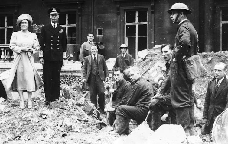 La foto en blanco y negro muestra que dos personas bien vestidas de pie junto a los trabajadores de rescate que están limpiando ruinas.