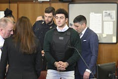 A man wearing a green sweater is handcuffed as he enters court with his lawyers.