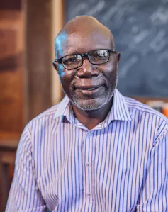 A bald-headed African man in glasses and a striped, collared shirt smiles at the camera.