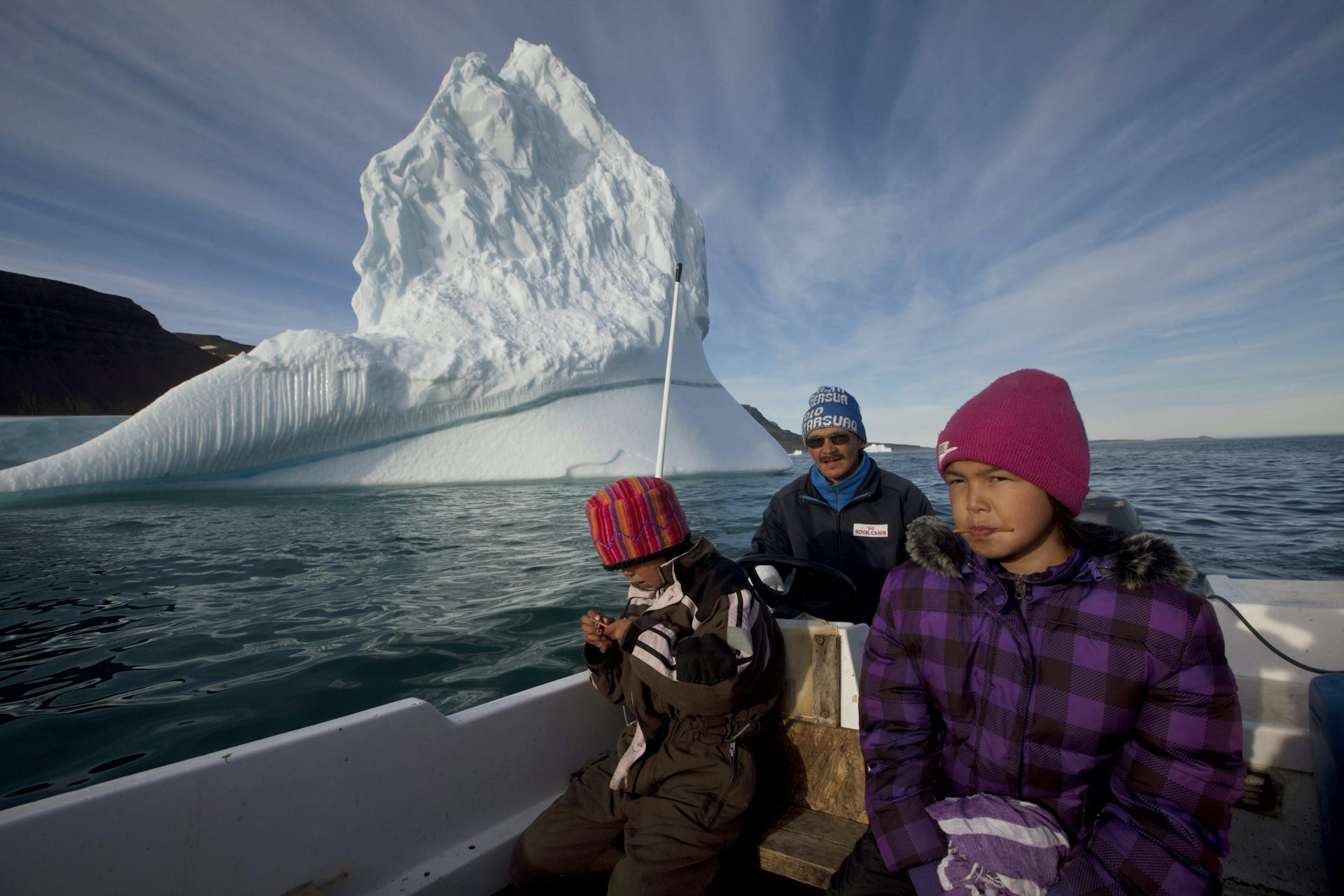 El hombre y dos niños sentados en un bote con un gran iceberg detrás de ellos.