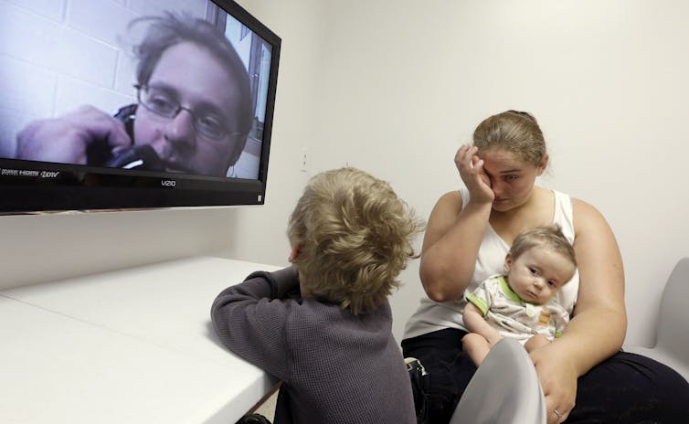 Woman holding baby rubs face and young child speaks to man on TV screen