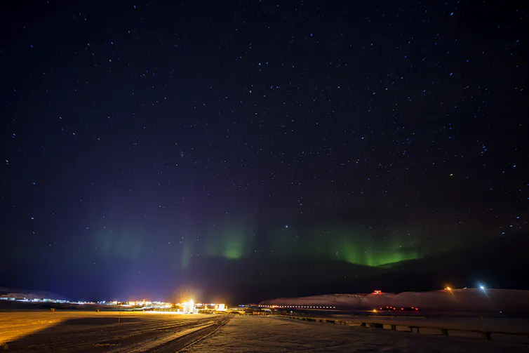 Several buildings on an icy plain, with green lights in the sky above.