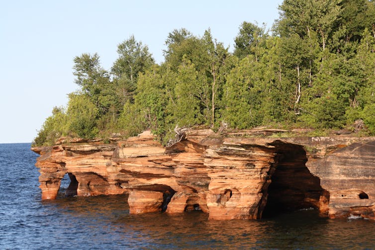 Cliffs with hollowed-out sections rise above blue water. Trees grow on the clifftops.