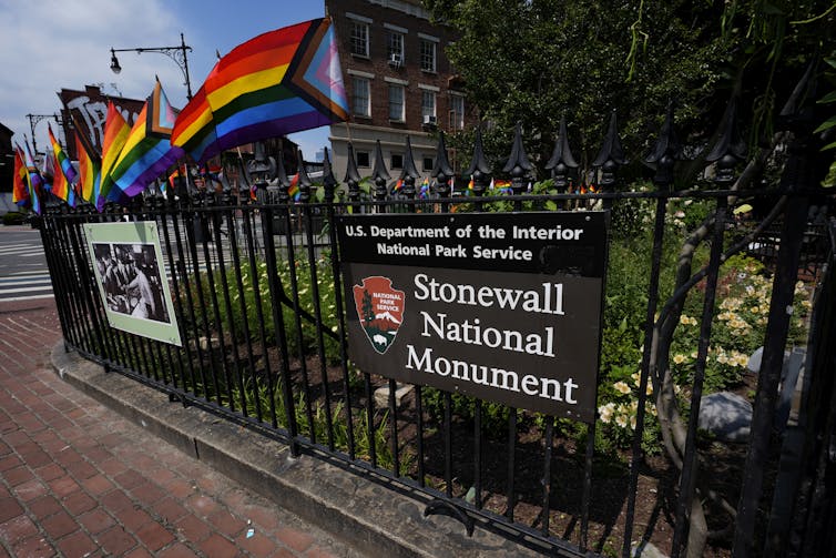 A sign reads 'Stonewall National Monument' next to a fence adorned with rainbow flags.